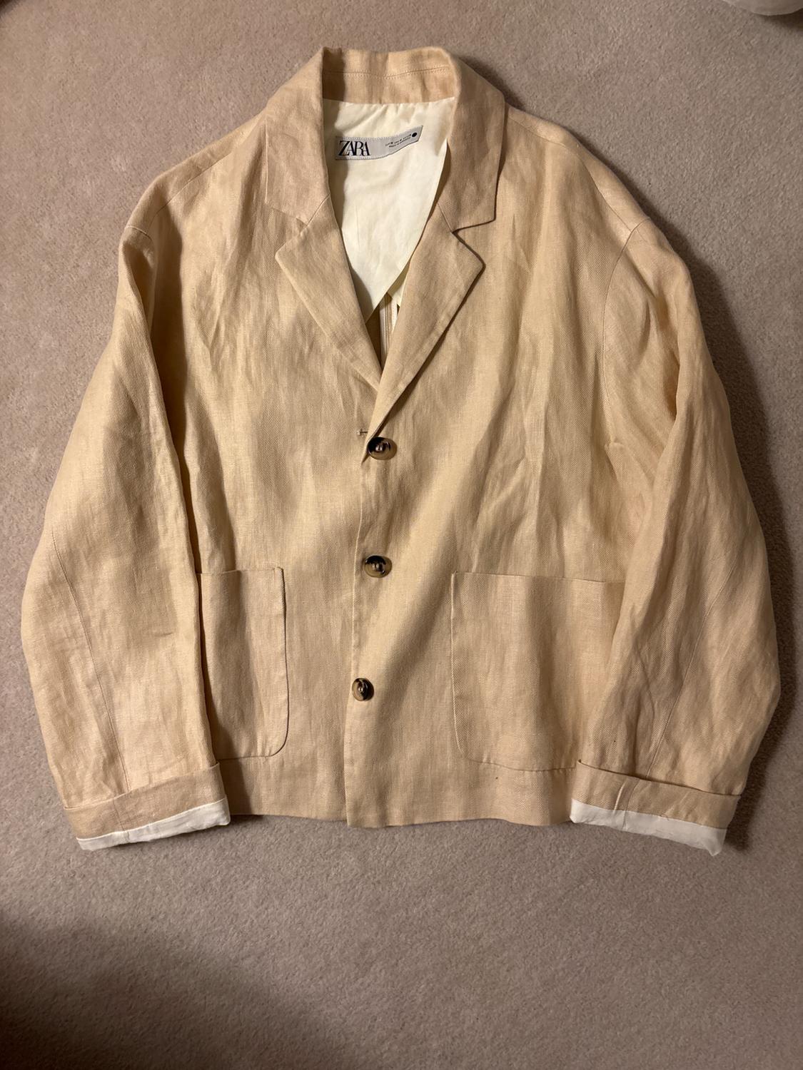 Beige linen jacket with three buttons and two front pockets, on a beige surface. Visible label: ZARA.