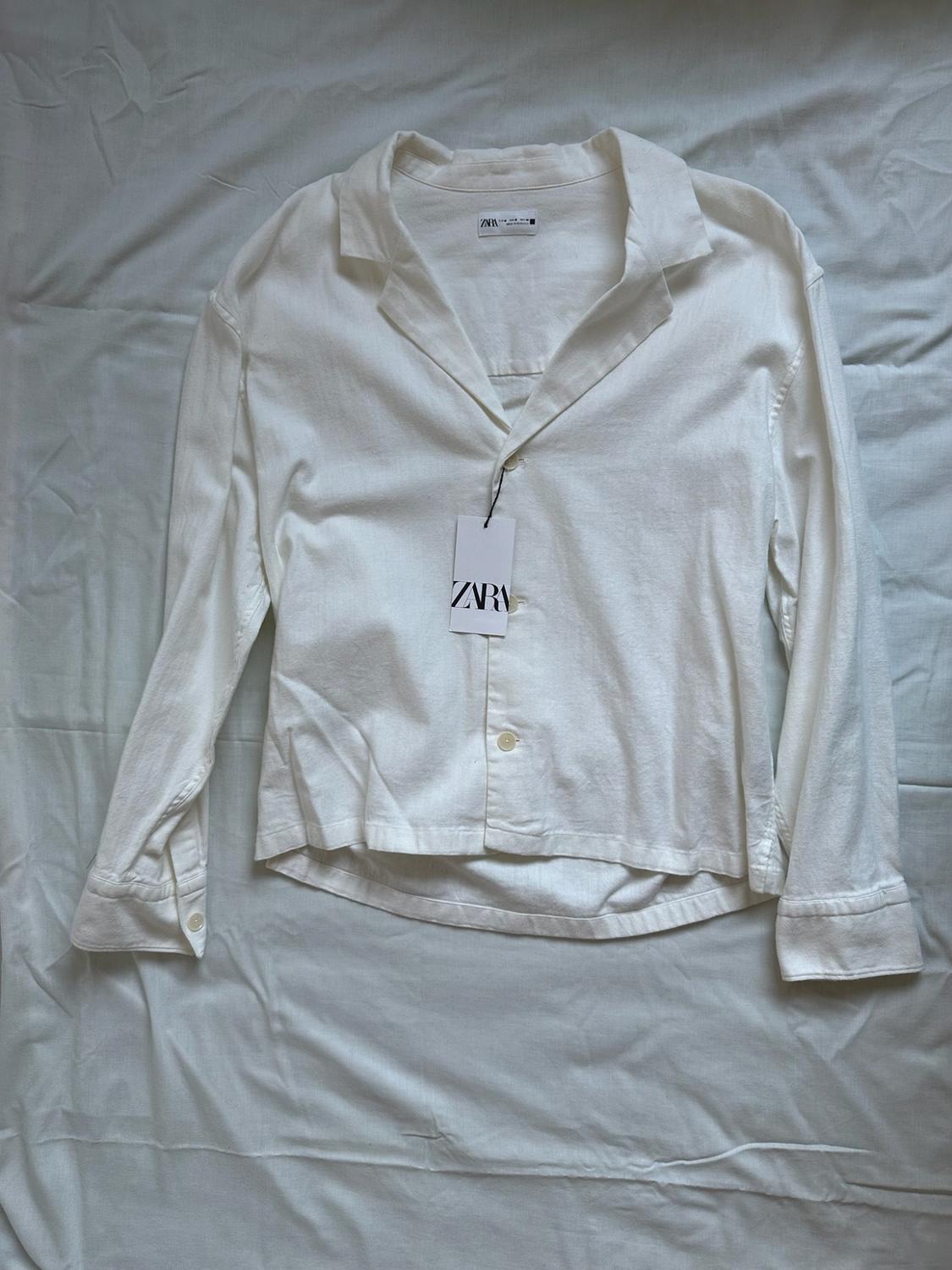 White long-sleeved shirt on a creased white sheet. Hanging tag with the text ZARA. Casual and relaxed style.