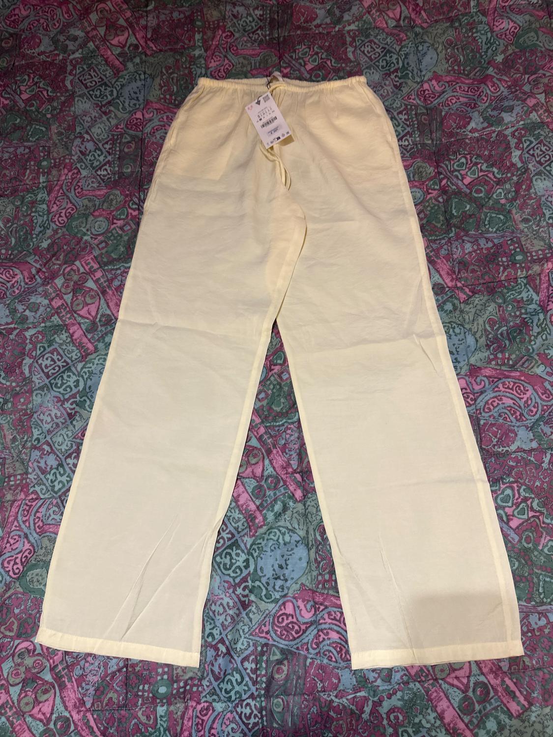 Light yellow trousers with a tag, on a background fabric with patterns in purple and green tones.