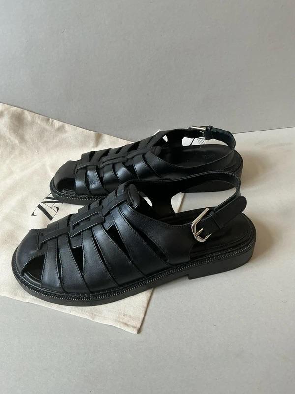 Black leather sandals with interwoven straps and a buckle, on a beige fabric bag with partially visible text.