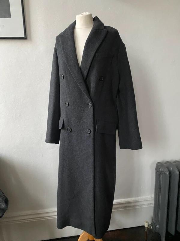 Dark grey, double-breasted long coat on a mannequin, in a room with white walls and a grey radiator in the background.