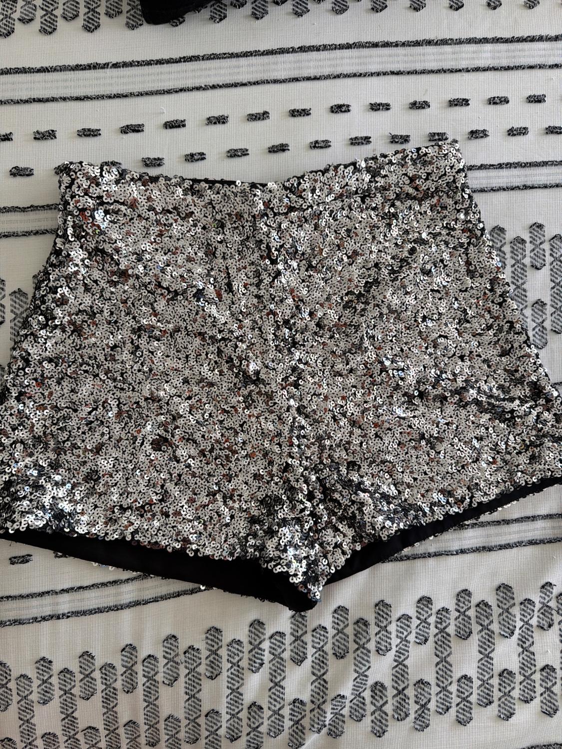 Silver sequinned shorts on a white fabric background with a black geometric pattern. Flat lay in a festive and relaxed style.