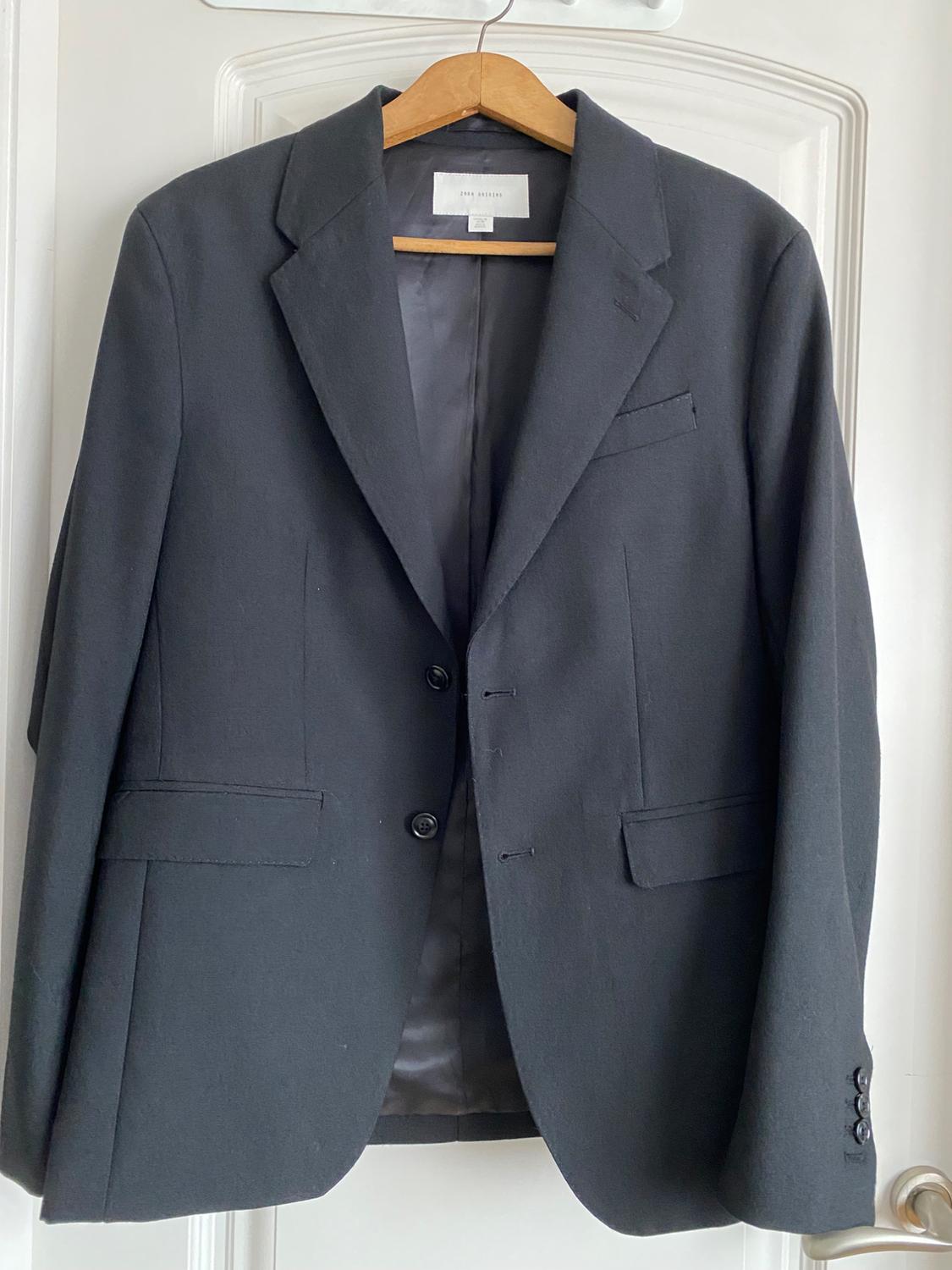 Black jacket hanging on a wooden hanger over a white door. Label visible on the inside of the collar.