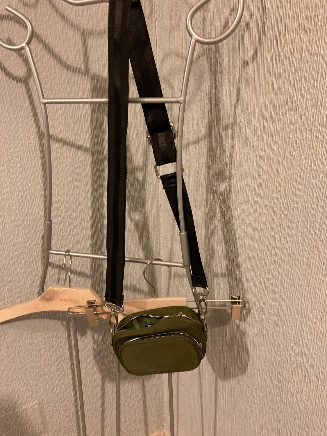 Olive green handbag with black straps hanging on a metal coat rack. Wooden hanger with "MANGO" text visible.