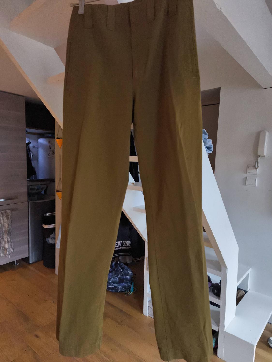Khaki trousers hanging on a hanger inside a room with a wooden floor and white stairs in the background.