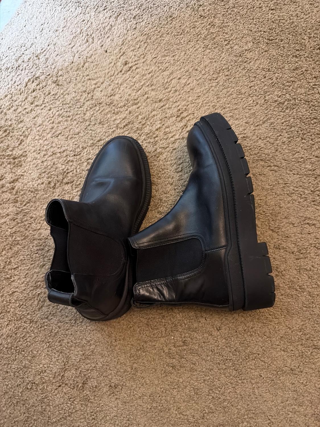Black leather boots with a chunky sole on a beige rug. Modern and robust design, ideal for an urban look.