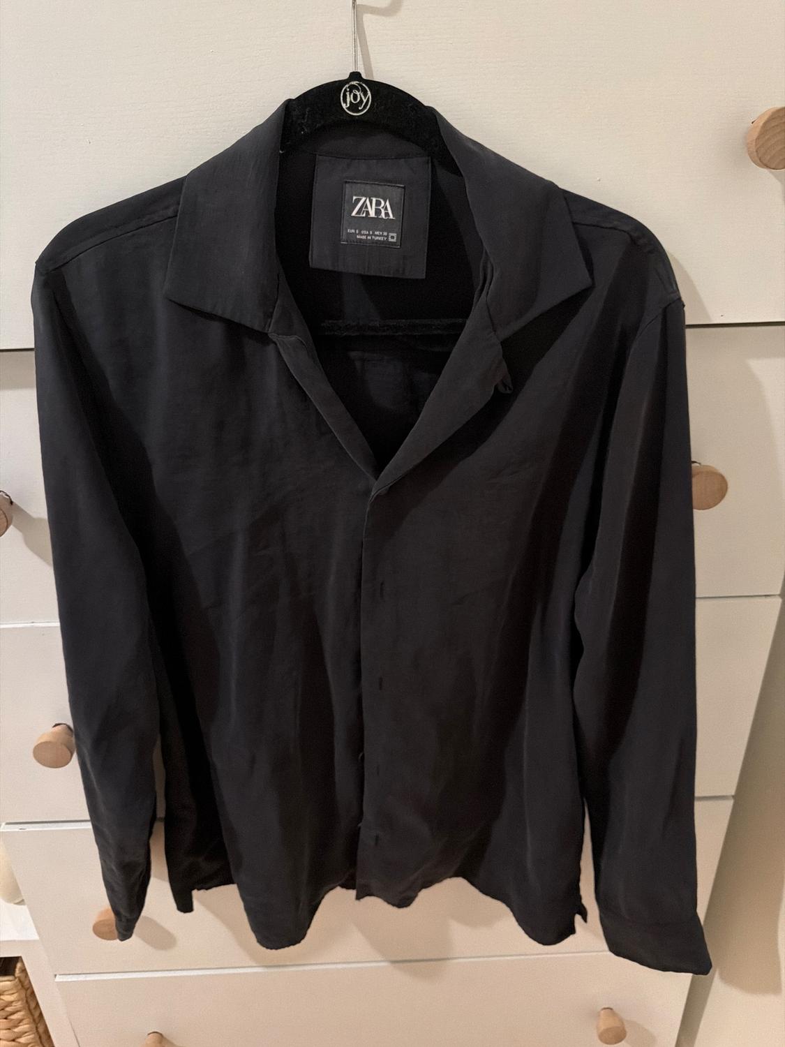 Black long-sleeved shirt hanging on a black hanger with the label "ZARA", against a white chest of drawers.