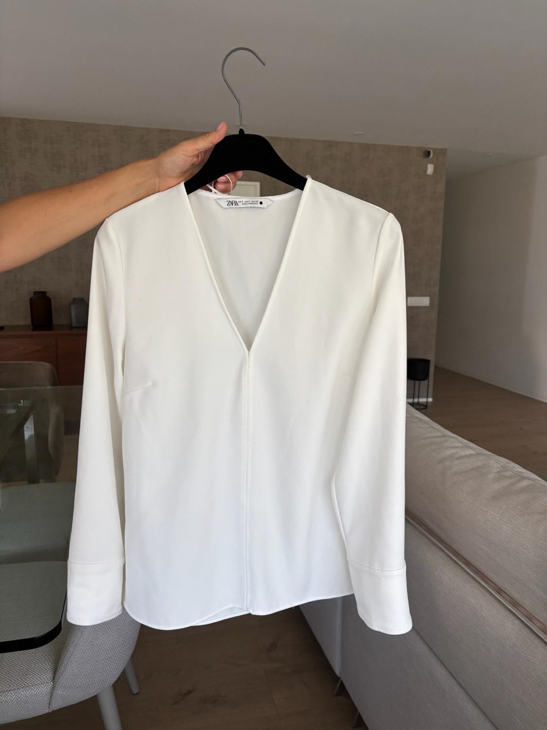 Hand holding a white long-sleeved V-neck blouse on a black hanger. ZARA label visible. Living room background with a grey sofa.