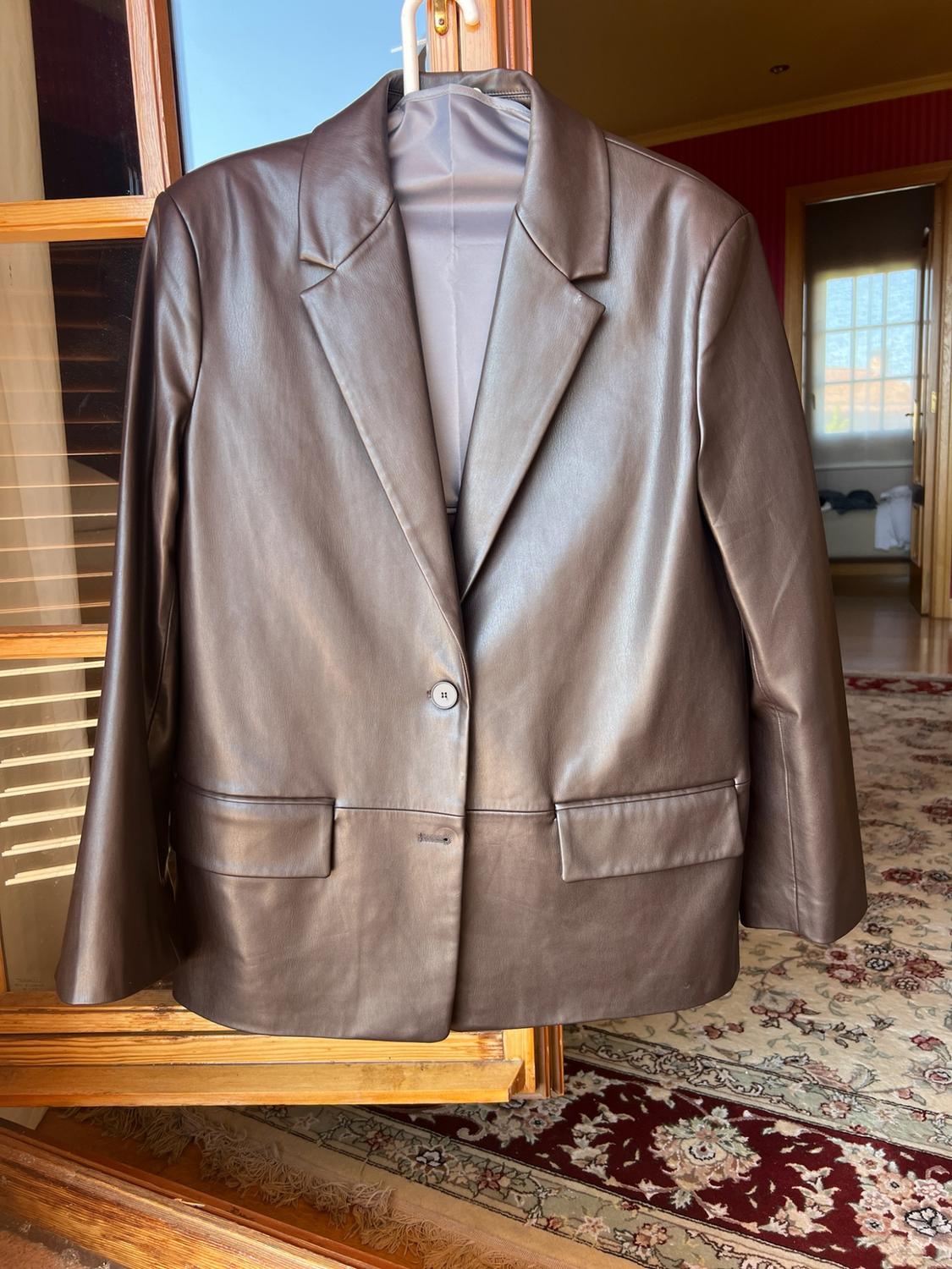 Brown single-button blazer with pockets, hanging on a wooden door. In the background, a room with a patterned carpet.