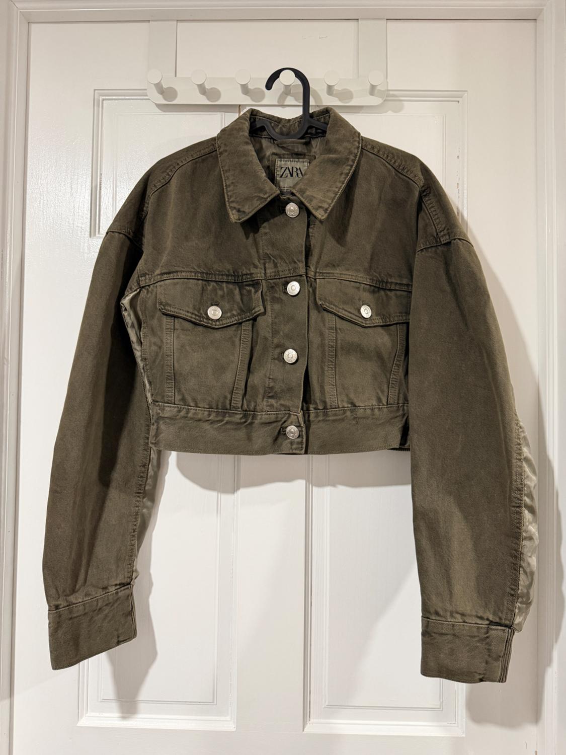 Olive green short jacket with silver buttons hanging on a black hanger over a white door. Visible label with text "ZARA".