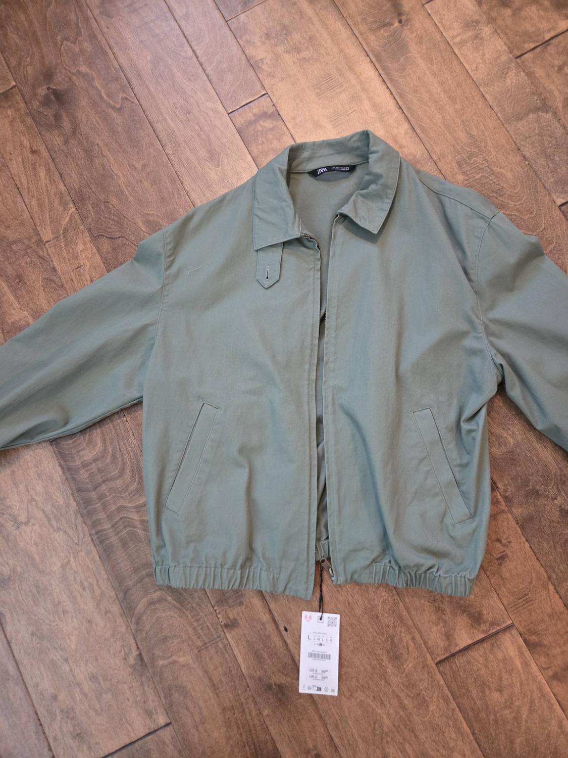 Sage green jacket with a collar and zipper, on a wooden floor. Includes a Zara label and a hanging price tag from the zipper.