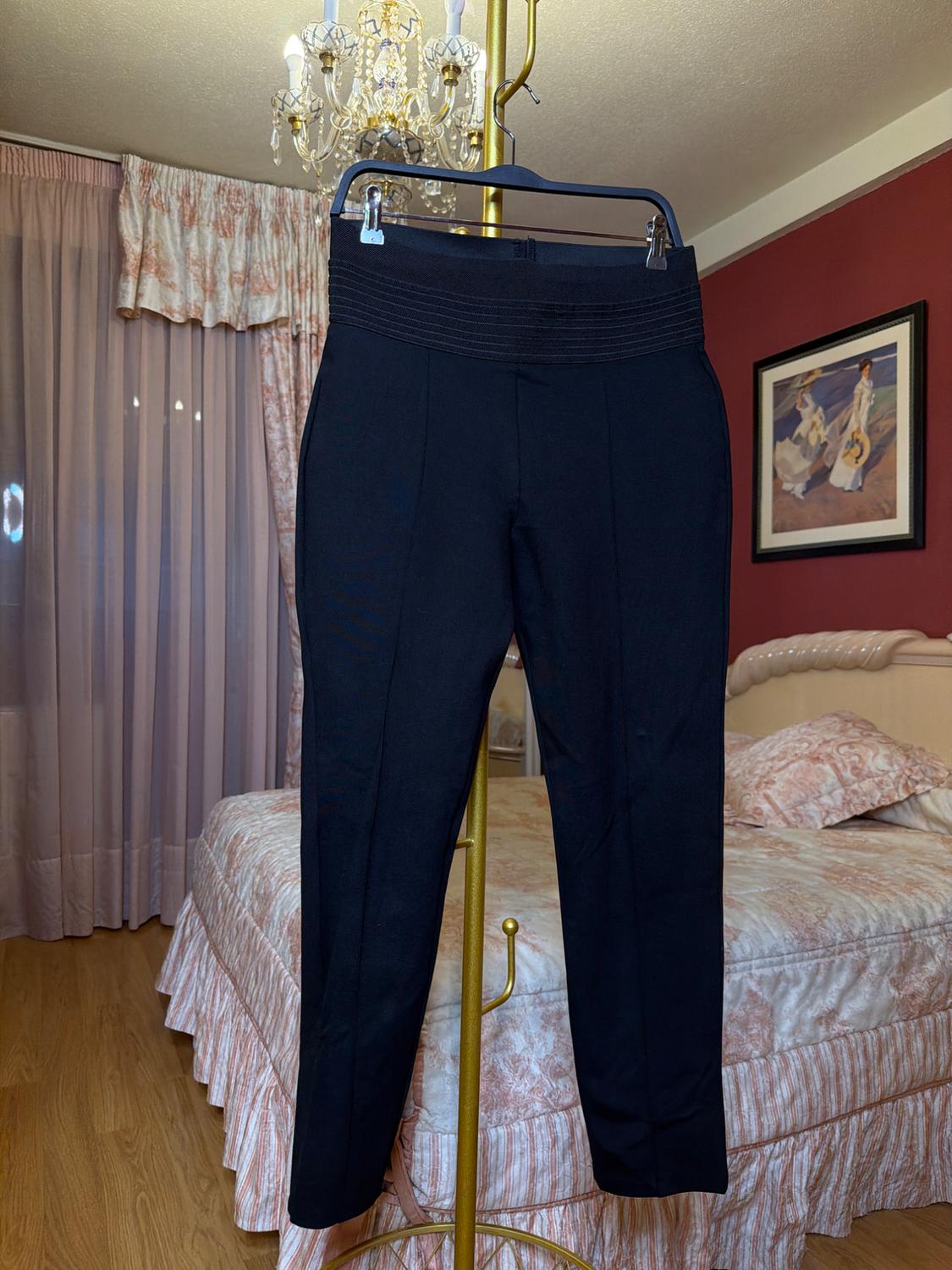 Black trousers hanging on a gold clothes rail in a bedroom with a patterned bedspread, curtains, and a chandelier.
