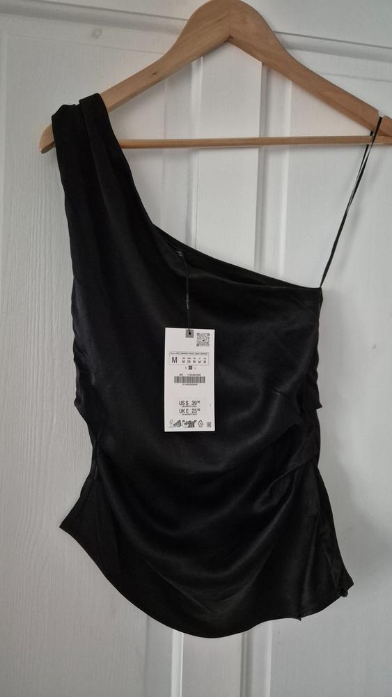 Black one-shoulder top hanging on a wooden hanger. Visible tag with price: US$ 39, UK£ 25. White door background.