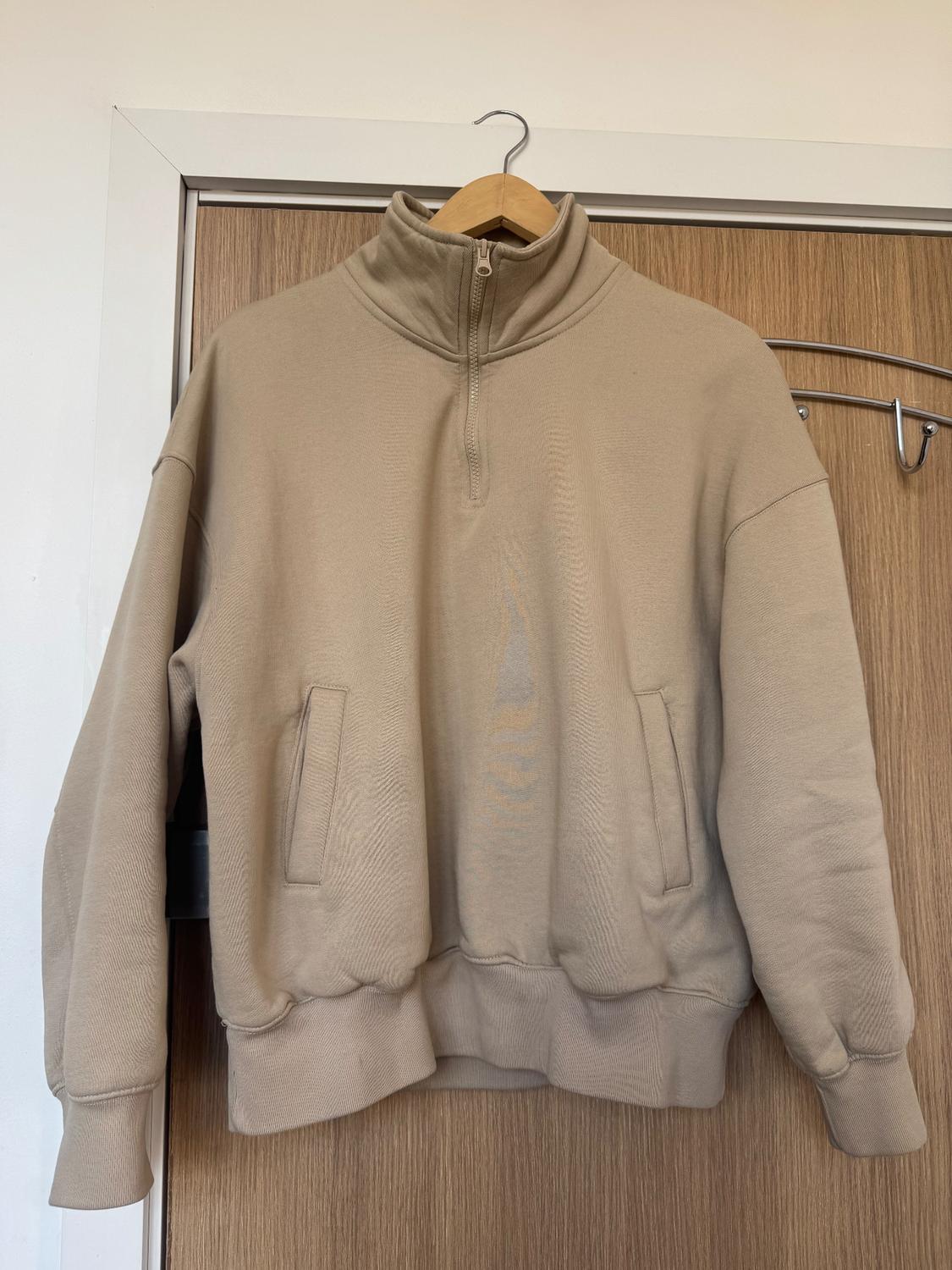 Beige sweatshirt with partial zip and high neck hanging on a wooden hanger over a light wood door.