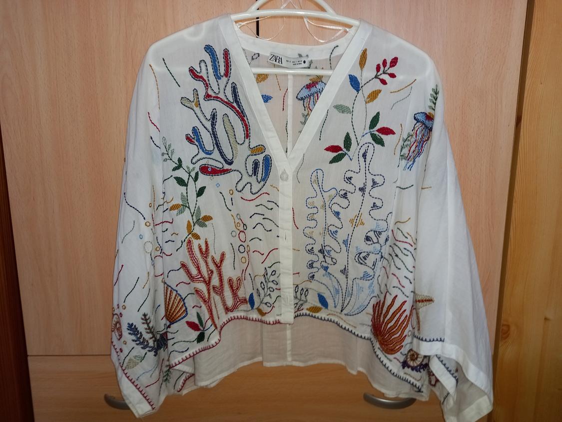 White blouse with colourful marine-themed embroidery, hanging on a light wooden hanger. Visible label: ZARA.