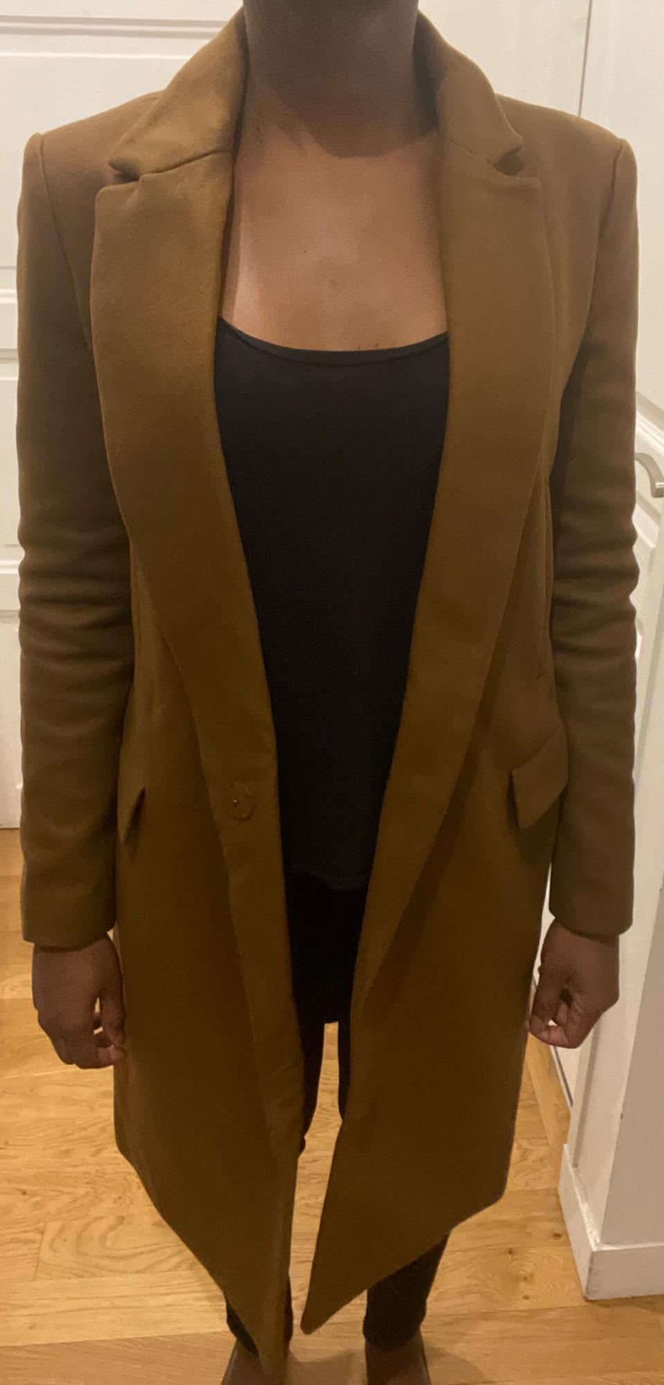 Model in a long brown coat over a black t-shirt, standing indoors with a wooden floor and white door in the background.