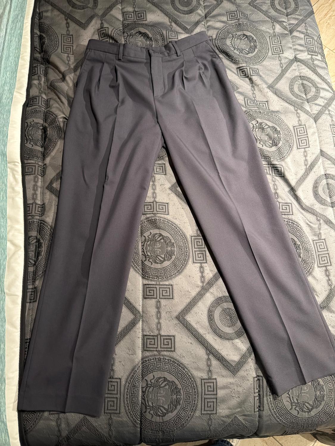 Dark grey trousers with pleats, laid out on a bedspread with a geometric design and circular patterns in shades of grey.