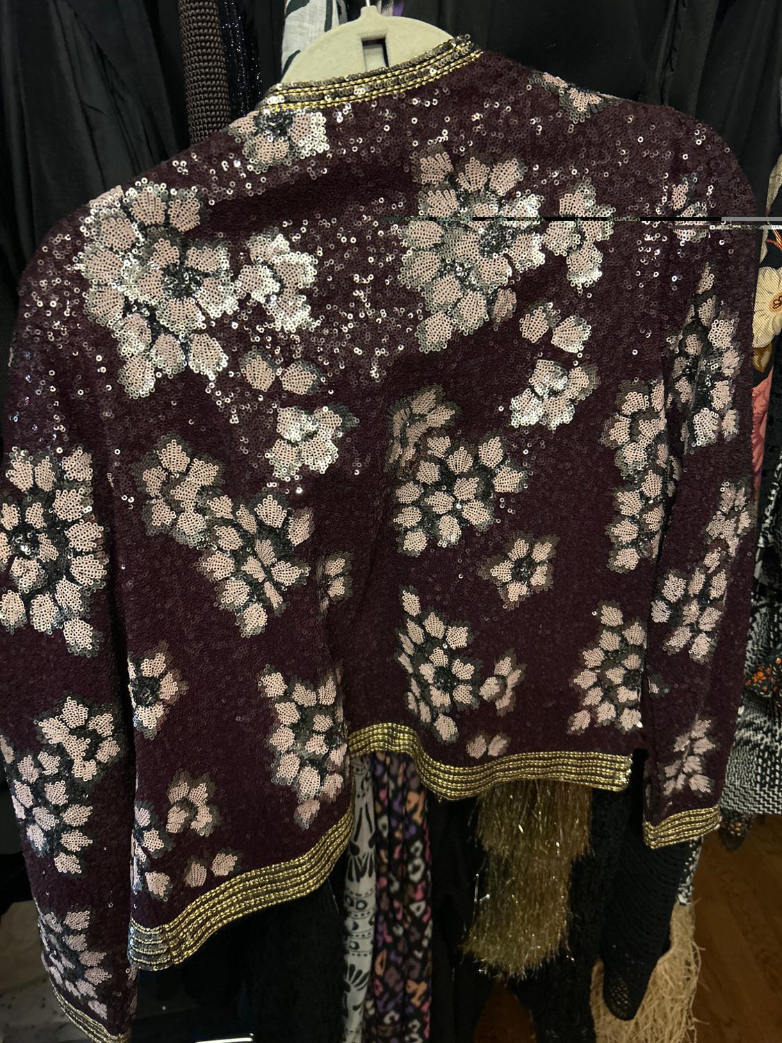 Burgundy sequin jacket with white floral print and gold detailing on the collar and hem, hanging on a coat rack.