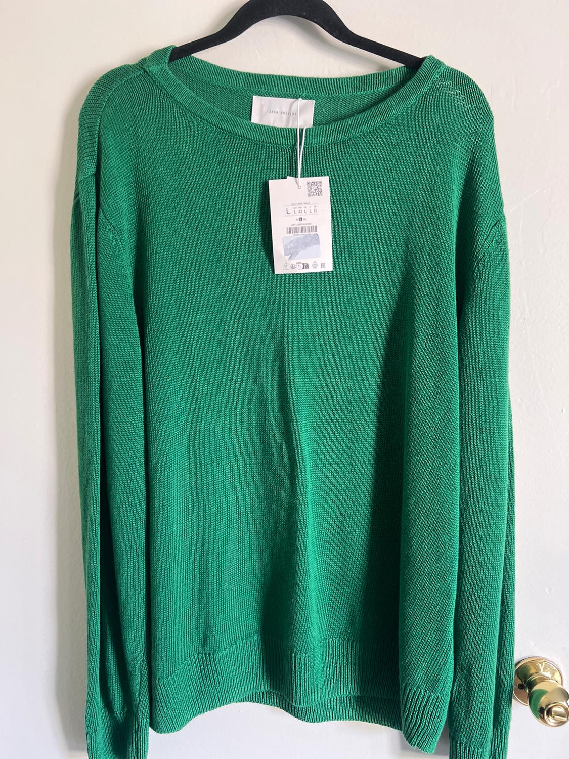 Green jumper hanging on a black hanger, with visible tag showing a QR code and text. Simple white background.