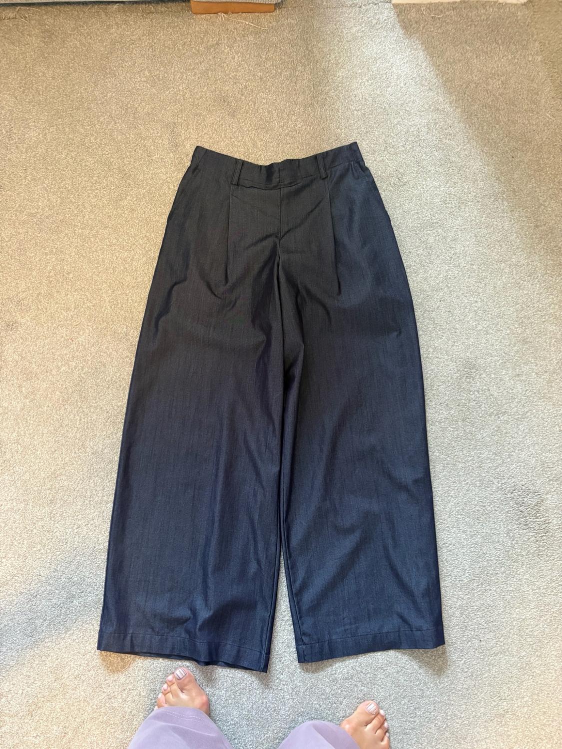 Dark blue wide-leg pleated trousers laid out on a grey carpet. Bare feet peep out from the bottom hem.