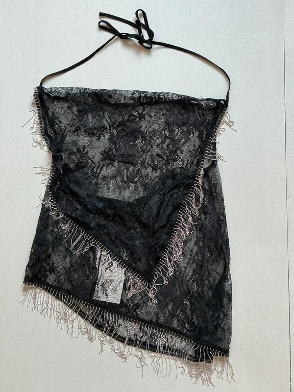 Black lace top with fringe and spaghetti straps on a white background. Transparent floral design and visible tag.