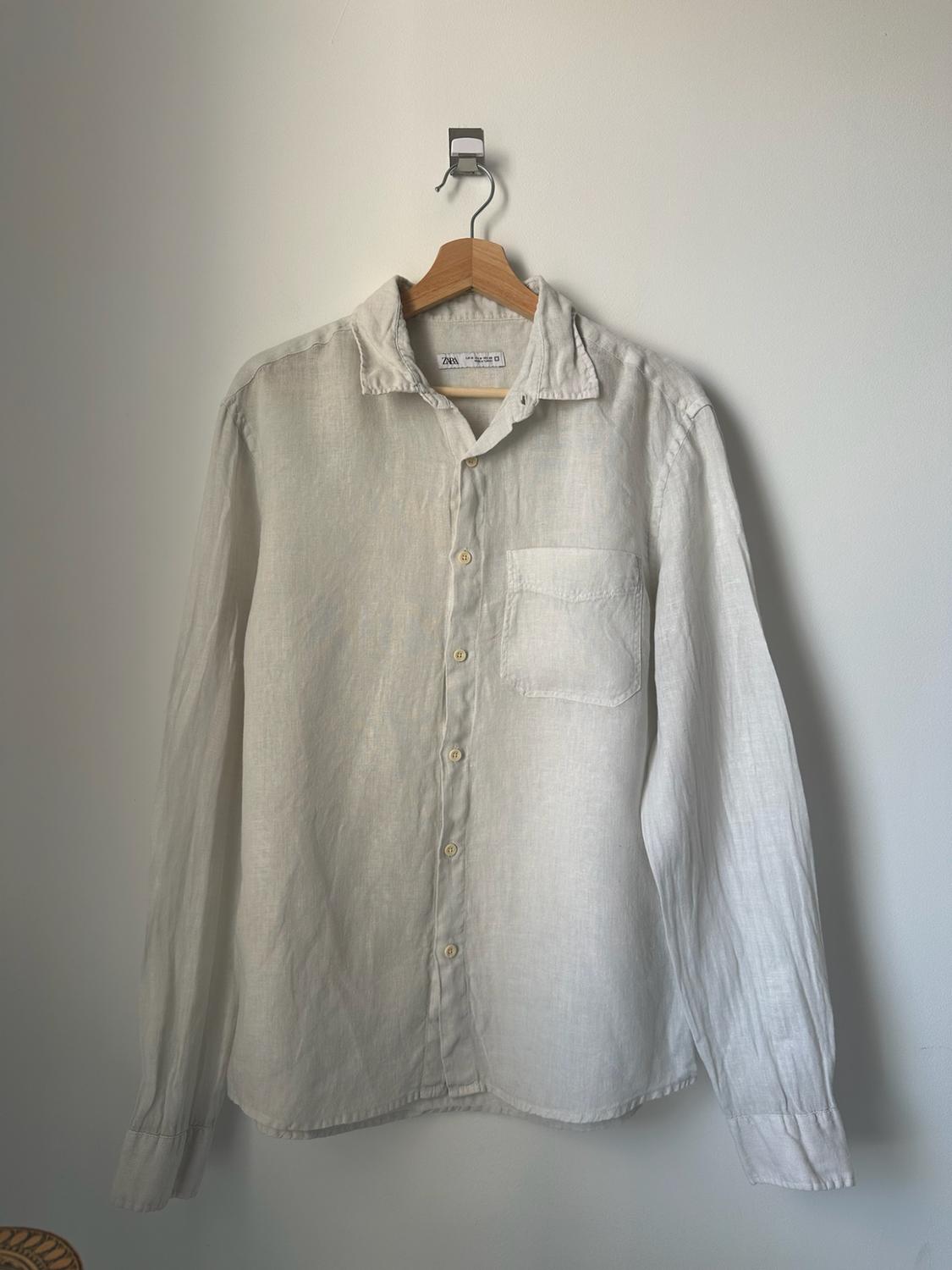 White long-sleeved button-up shirt with a pocket, hanging on a wooden hanger against a white wall. Text: ZARA.