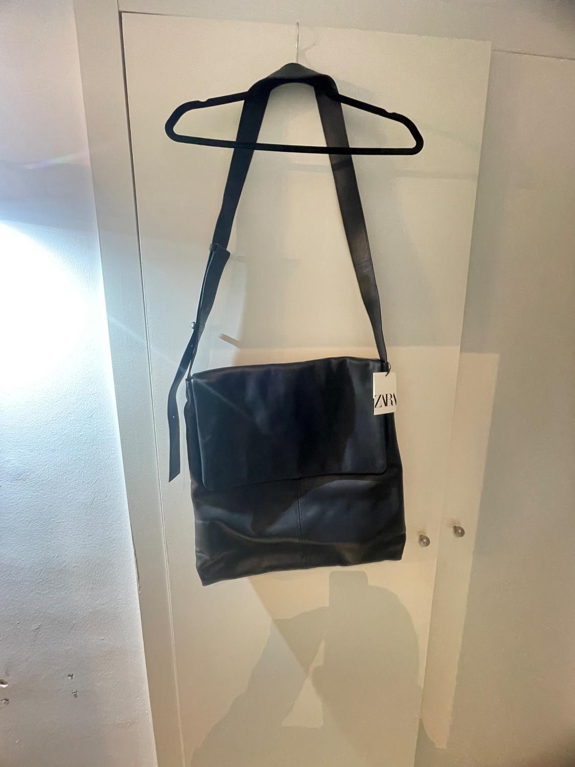 Black handbag hanging on a black hanger over a white door. Visible tag with the text "ZARA". Soft light to the left.