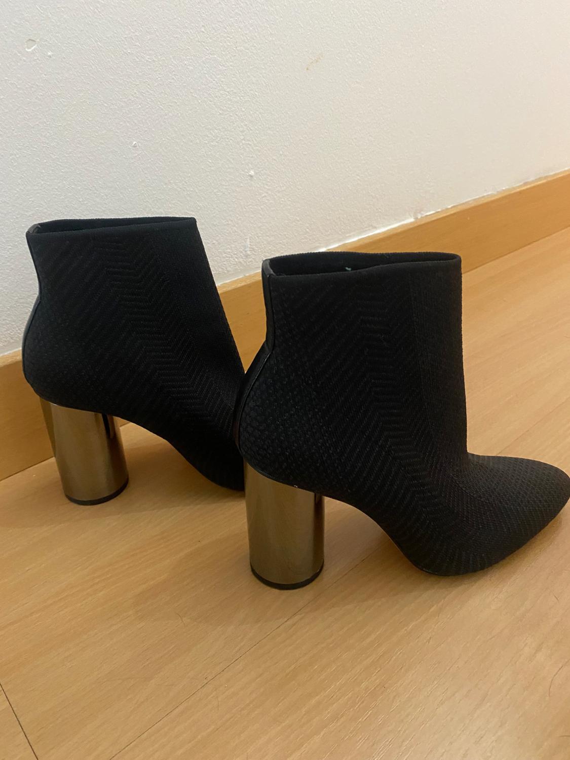 Black ankle boots with a herringbone texture and chunky metallic heel on light wooden flooring. White wall background.