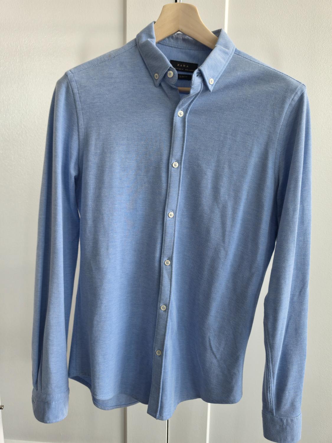 Light blue long-sleeve casual shirt, hanging on a wooden hanger in front of a white door. Label with the text ZARA.