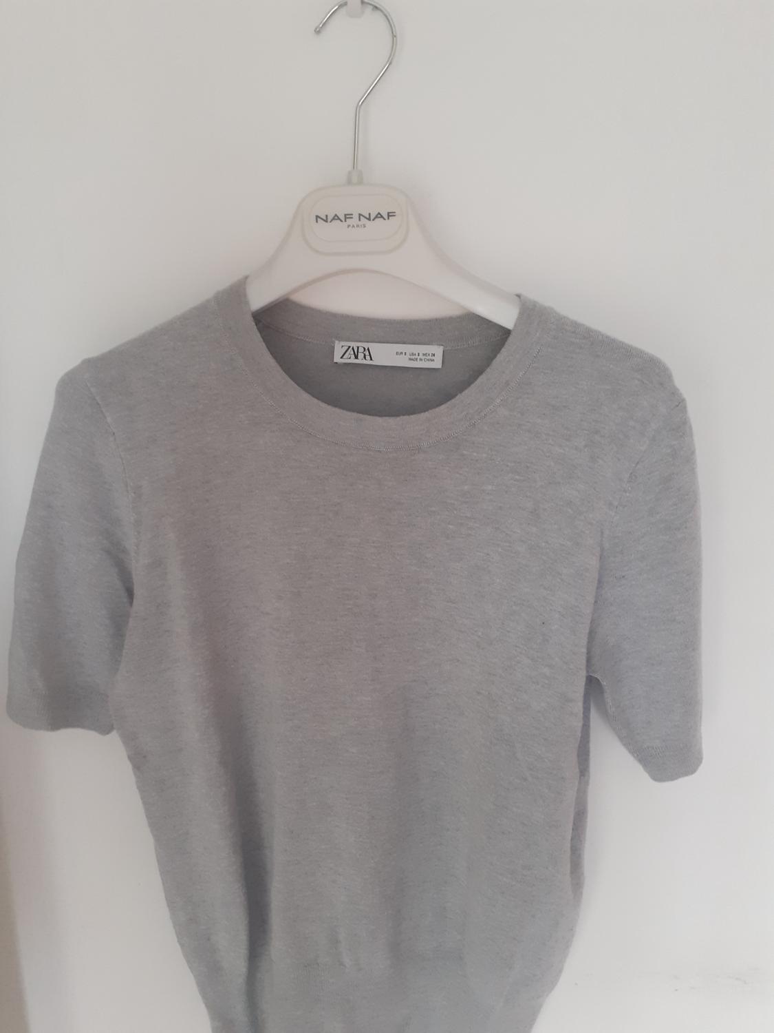 Grey short-sleeved knitted top on a white hanger with NAF NAF PARIS text. ZARA label on the neck. White background.