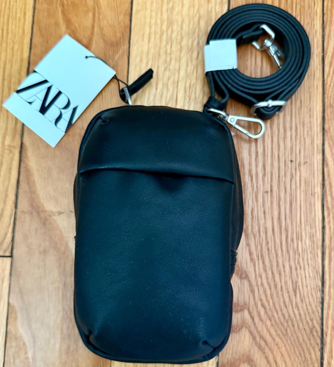 Black handbag with adjustable strap and Zara tag on wooden floor.