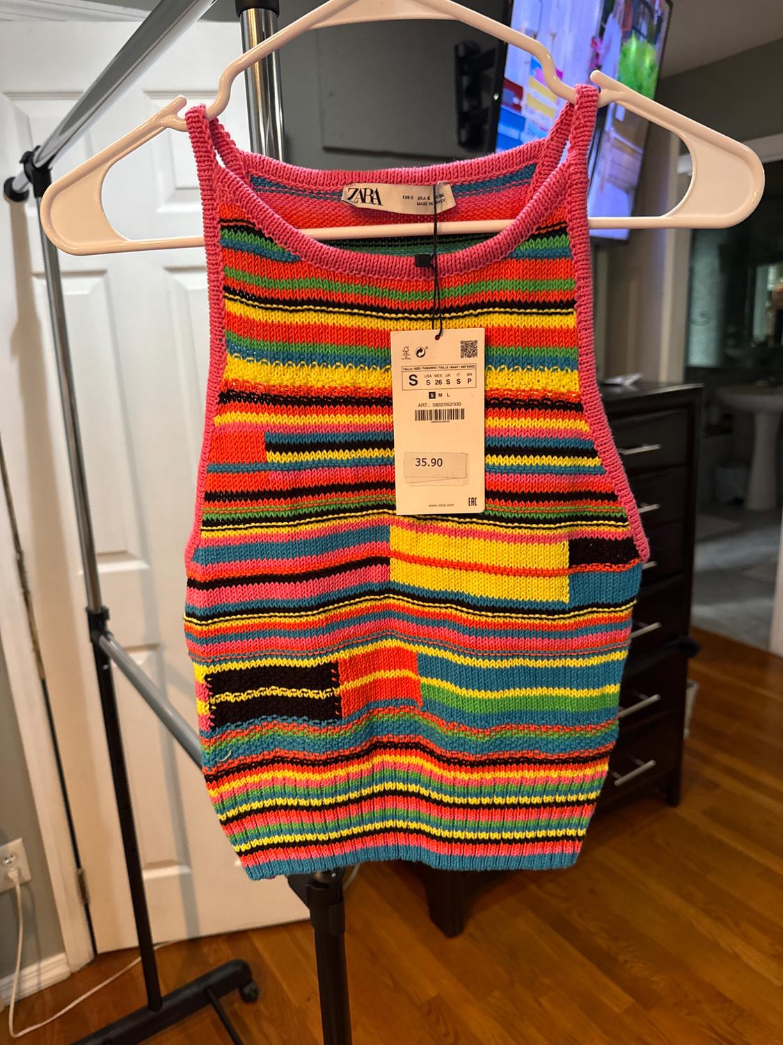 Multicoloured knitted top with stripes and a price tag hanging, hung on a white hanger against an indoor background.