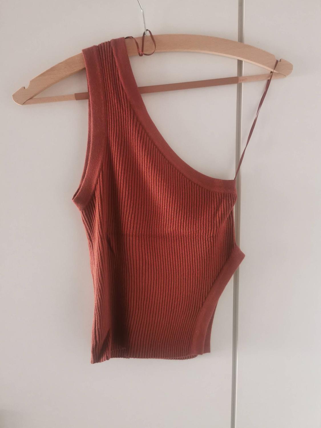 Terracotta ribbed knit one-shoulder asymmetric top, hanging on a wooden hanger in front of a white wardrobe.