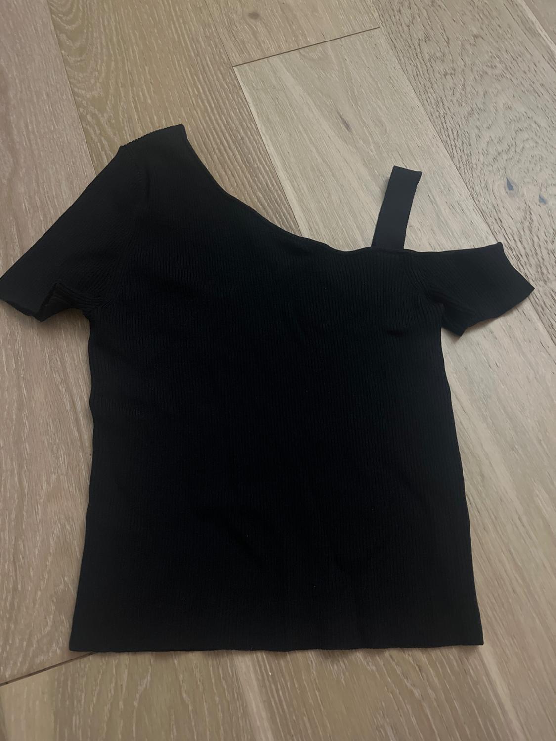 Black one-shoulder top on light wood floor. Asymmetrical design with a strap on the right shoulder.