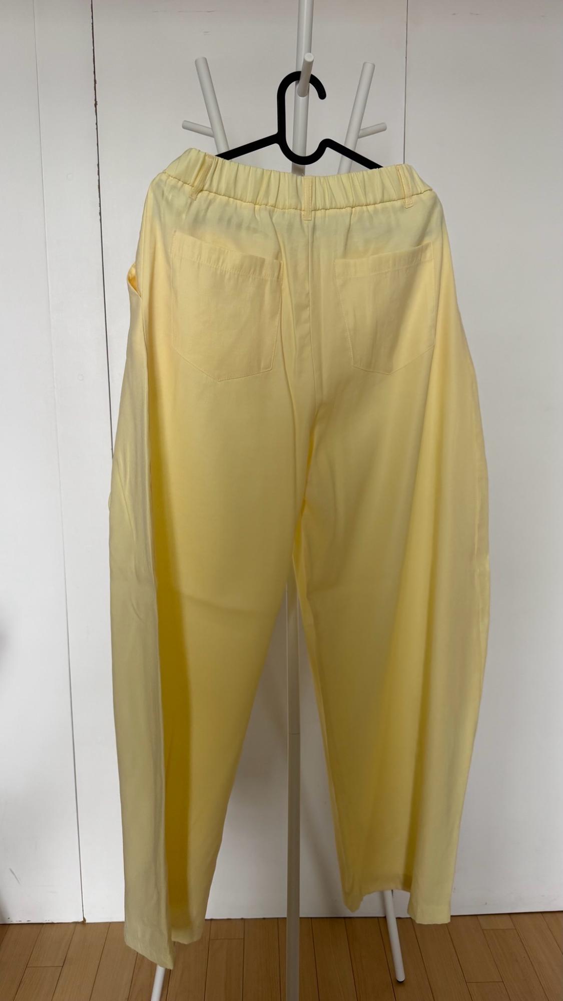 Light yellow trousers with an elasticated waistband and back pockets, hanging on a black hanger on a white clothes rack. White door background.