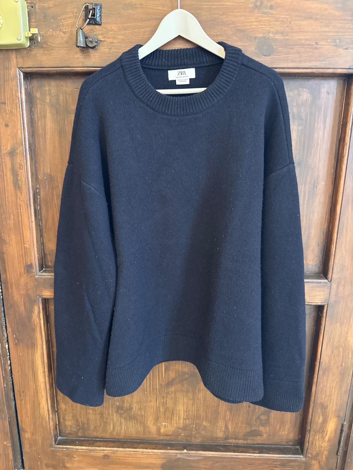 Black knitted jumper hanging on a white hanger over a wooden door. Visible label with the text "ZARA".