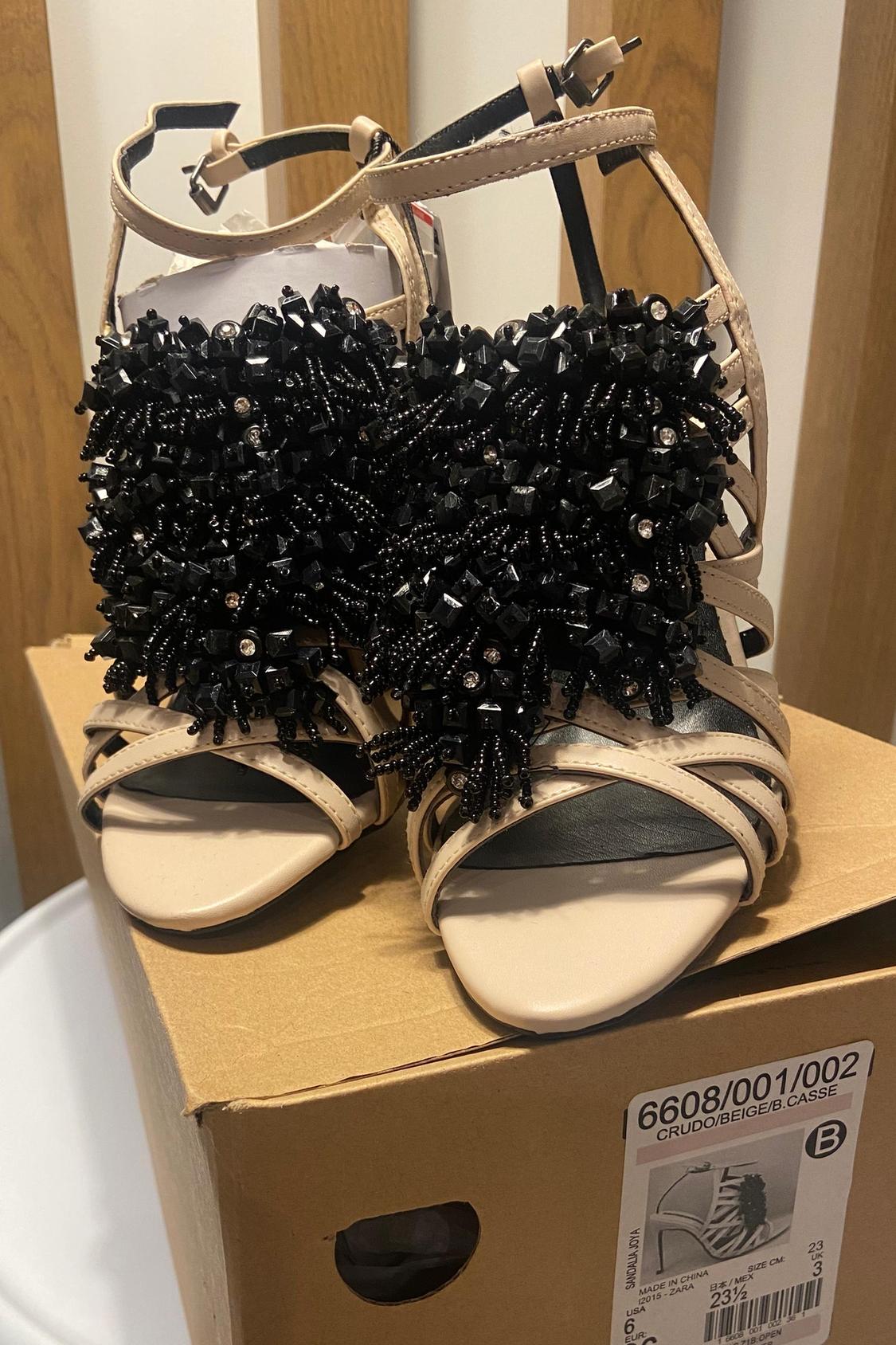 Beige sandals with black bead embellishments in a cardboard box.