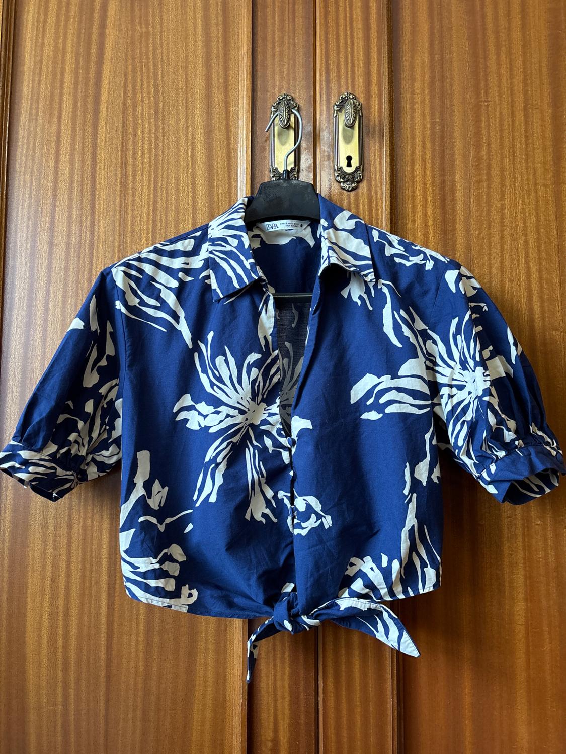 Blue shirt with white floral print, short sleeves and a knot at the waist, hanging on a hanger in front of a wooden door.