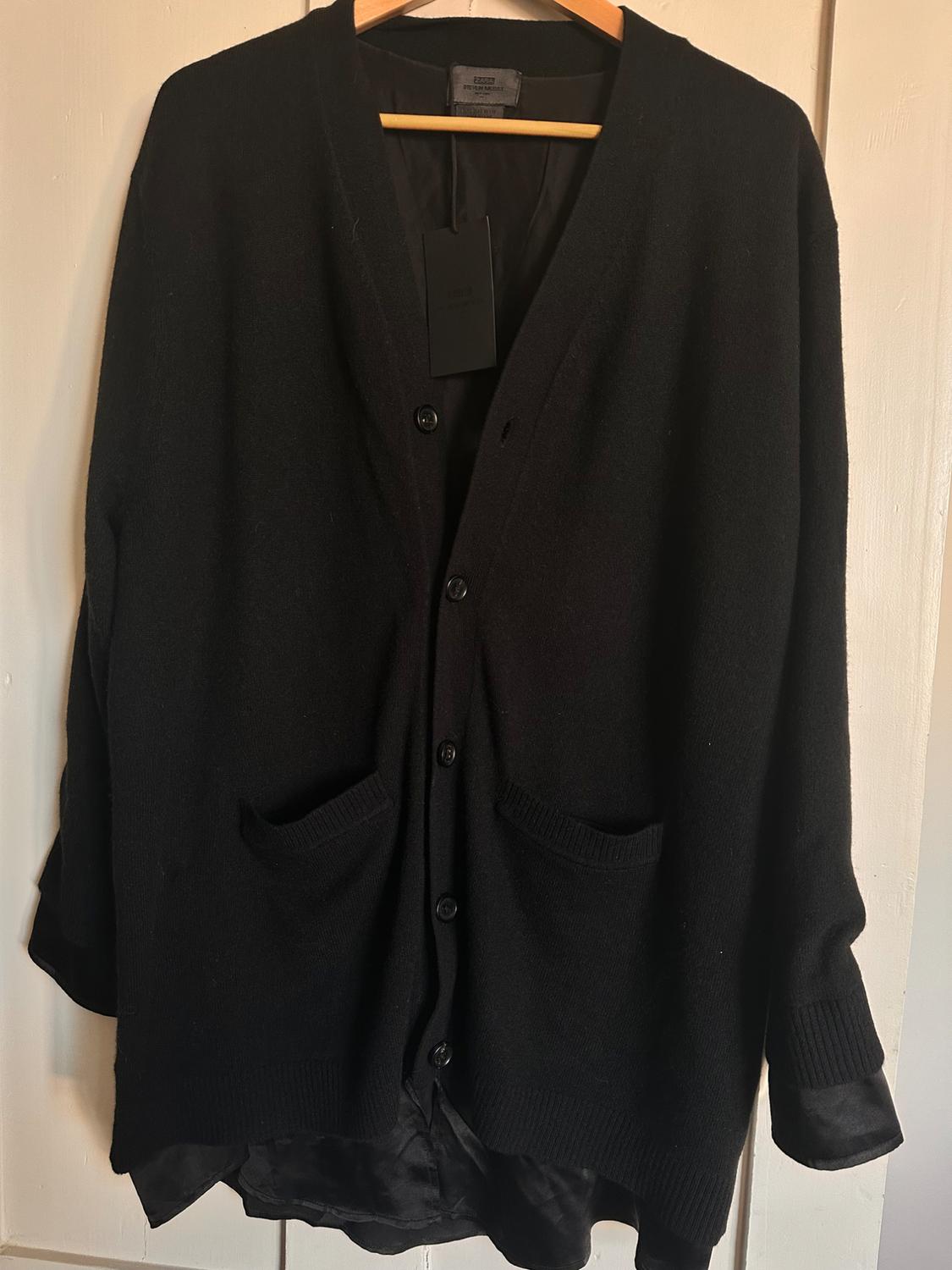 Black knitted cardigan hanging on a wooden hanger, with buttons and two front pockets. Black label visible on the inside.