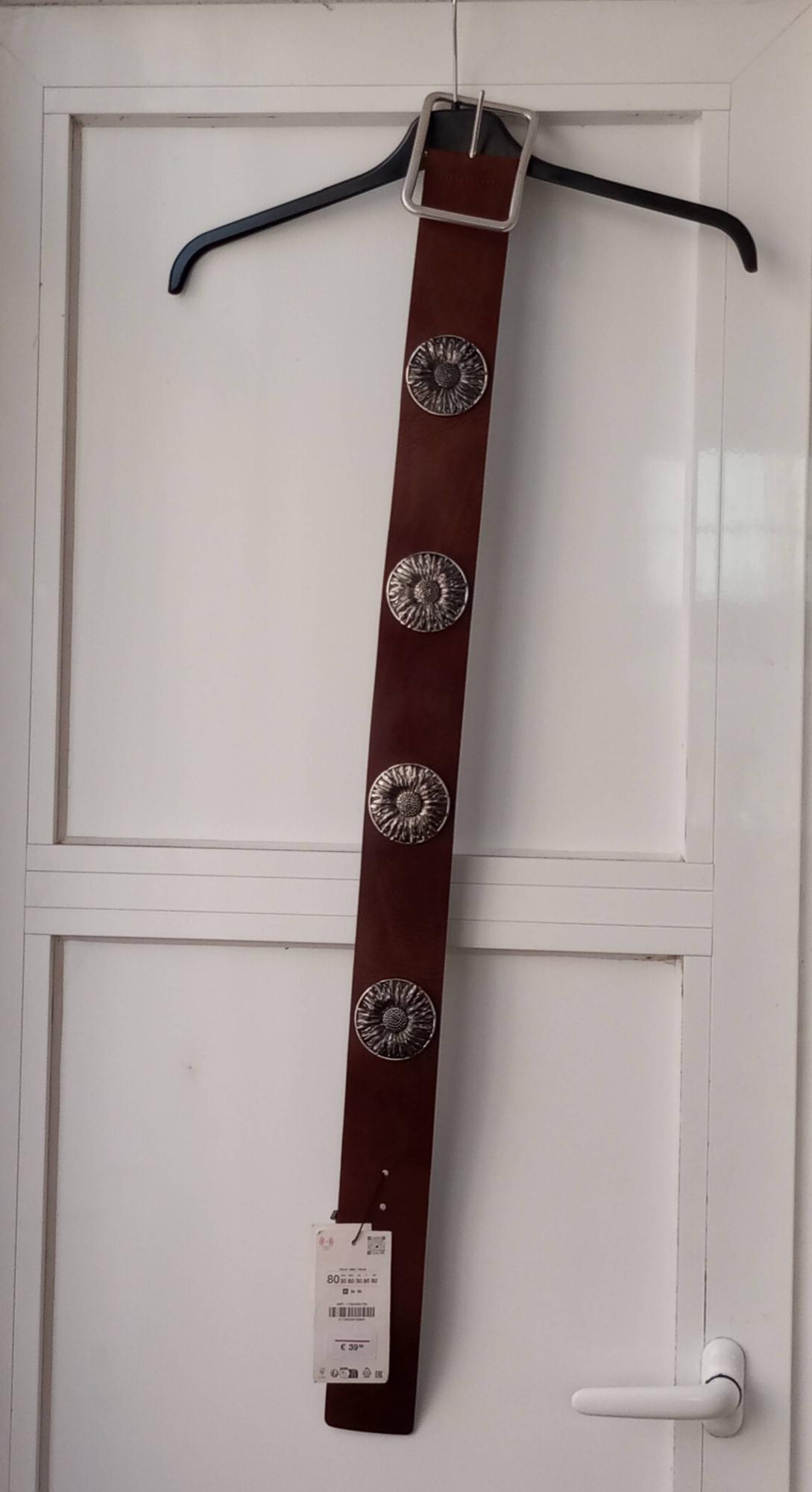 Wide brown belt with a square buckle and four large silver floral appliqués, hanging on a white door. Text: 80, €39.