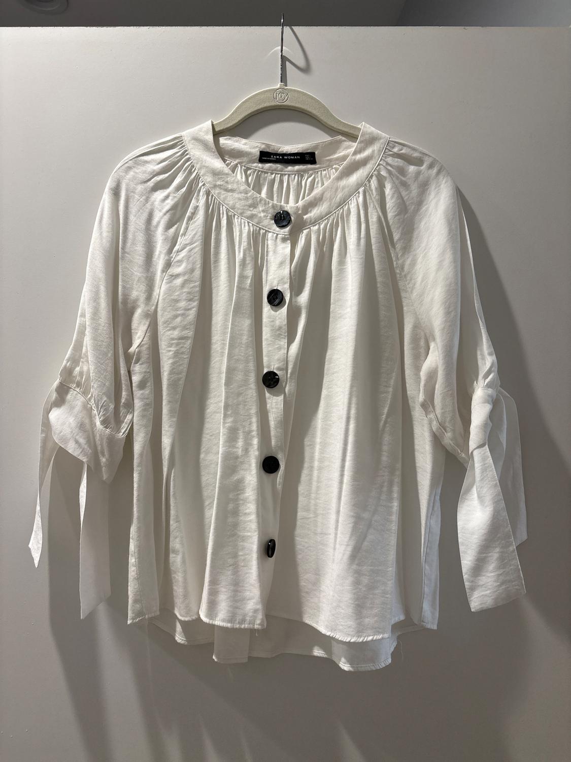 White long-sleeved blouse with black buttons, hanging on a hanger. Lightweight fabric with gathered detailing at the neck.