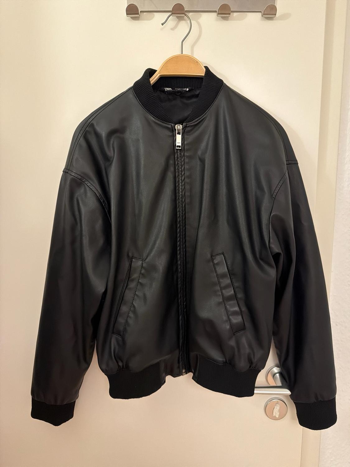 Black bomber jacket with a zip and elasticated trims, hanging on a wooden hanger over a white door.