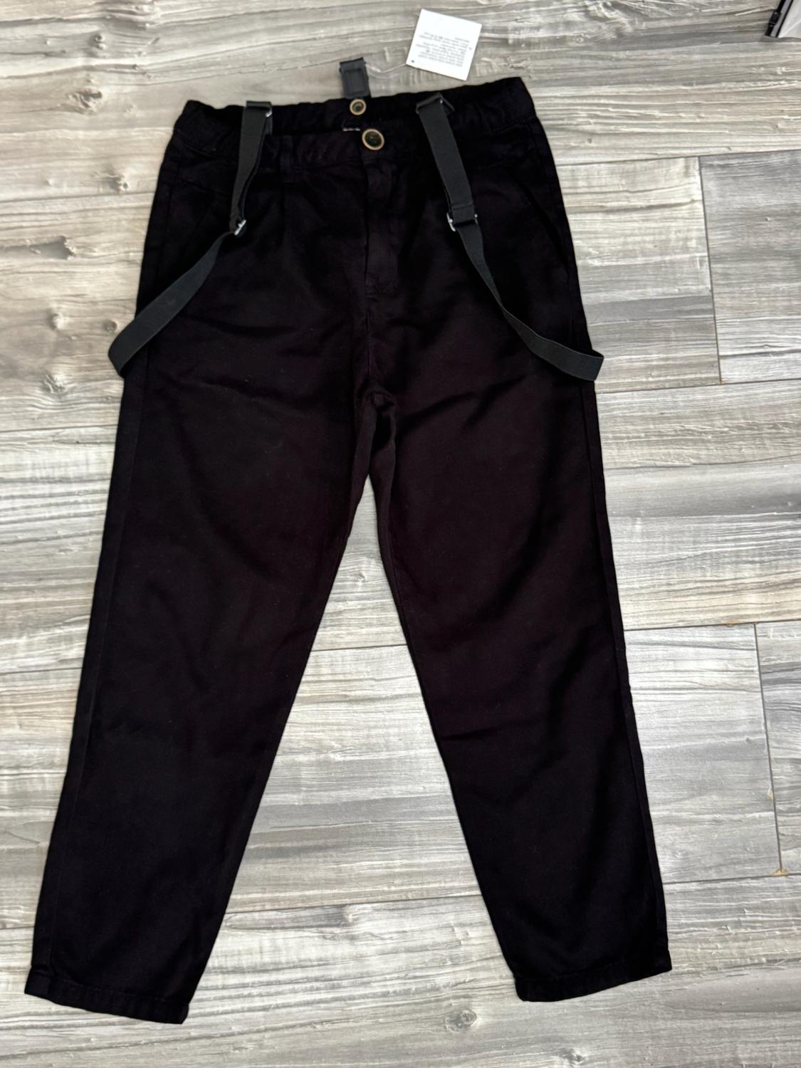 Black trousers with braces laid out on a grey wooden floor.