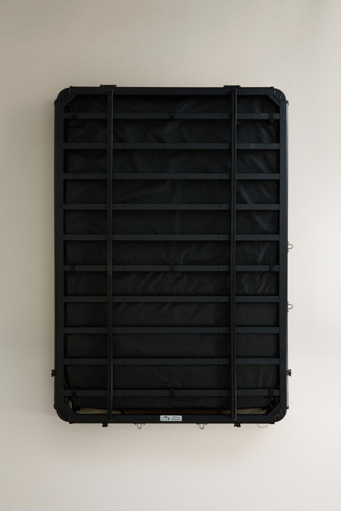 Front view of a folded roof tent with a black rectangular frame made of horizontal and vertical bars, and a label at the bottom.
