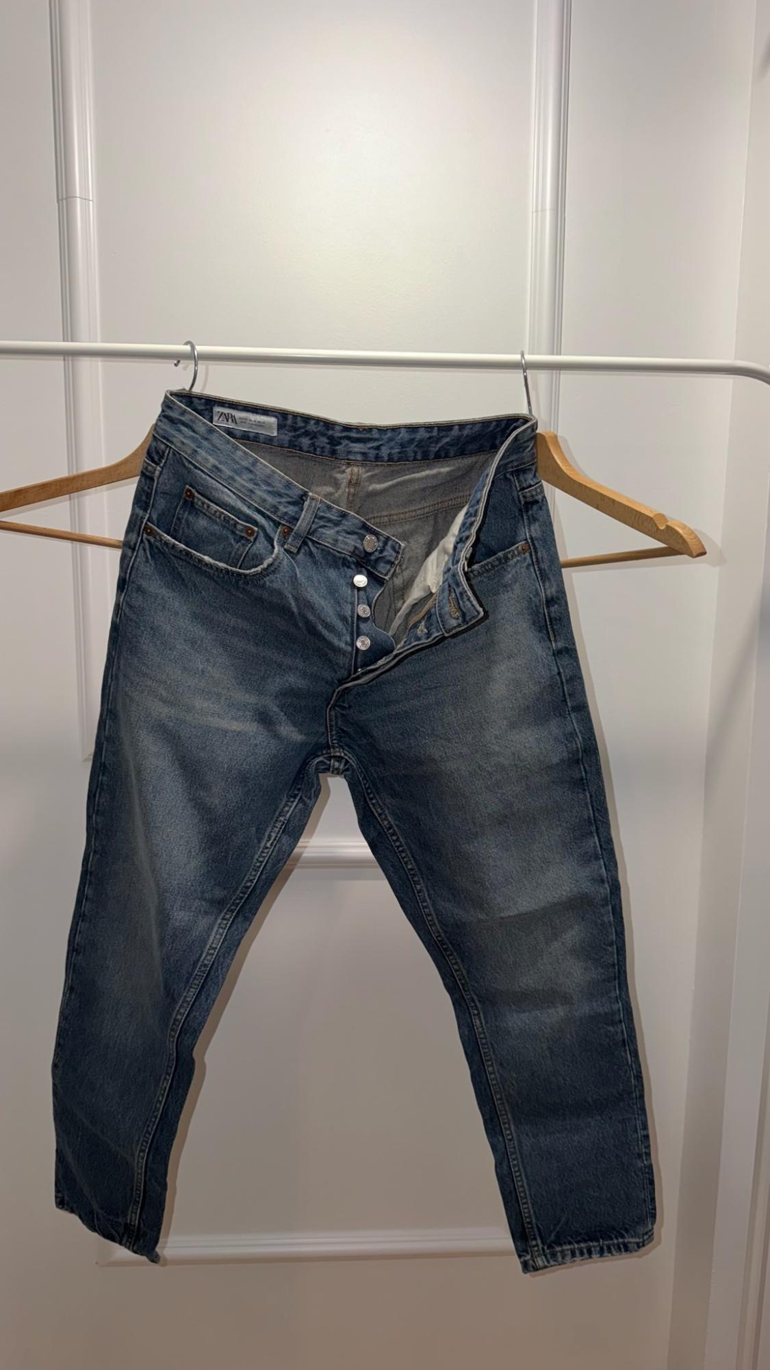 Unbuttoned blue jeans on a wooden hanger in front of a white door. The inner label reads ZARA.