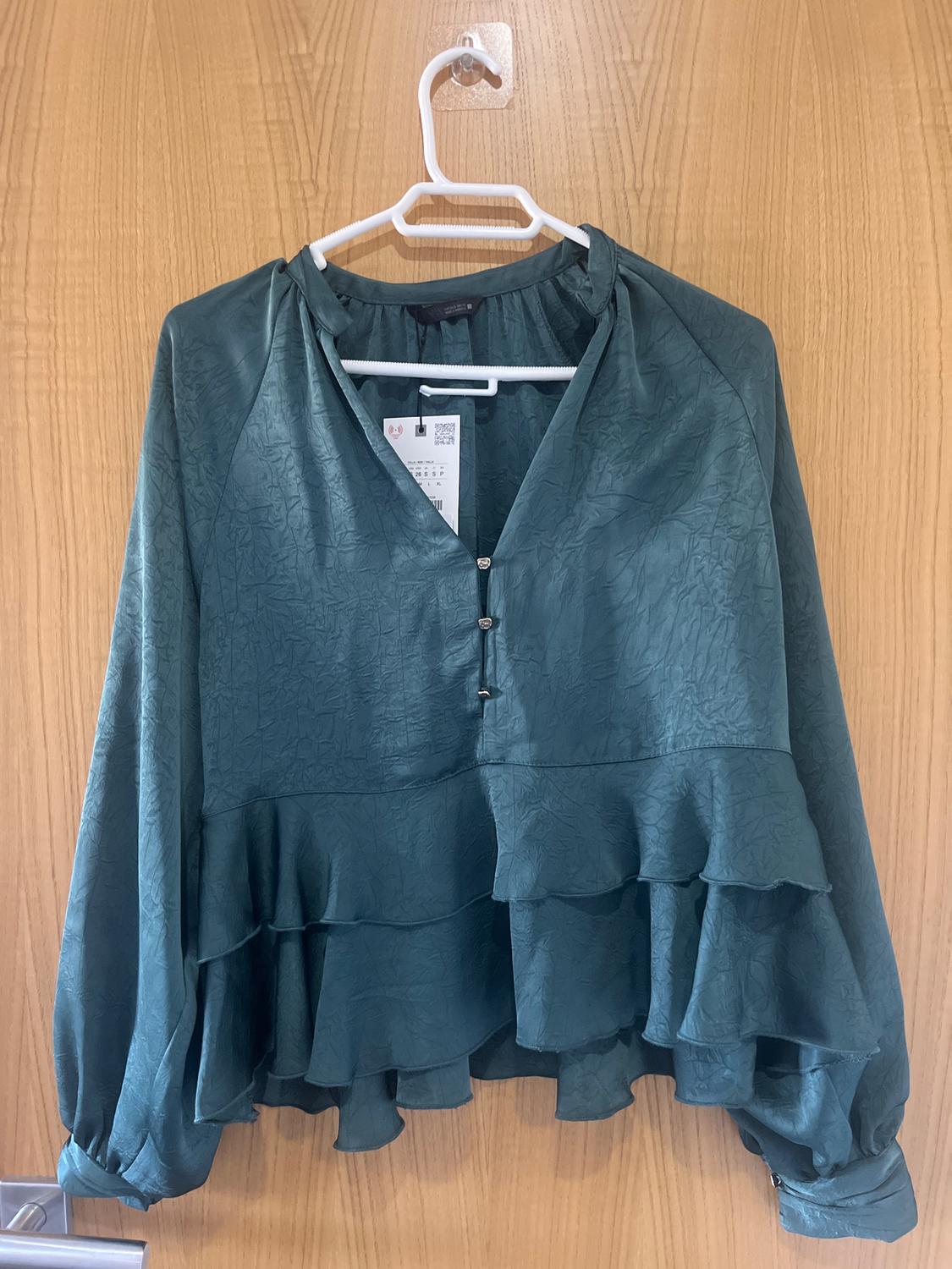 Green blouse with ruffles and a crinkled texture, hanging on a white hanger over a light wooden door. Label visible.