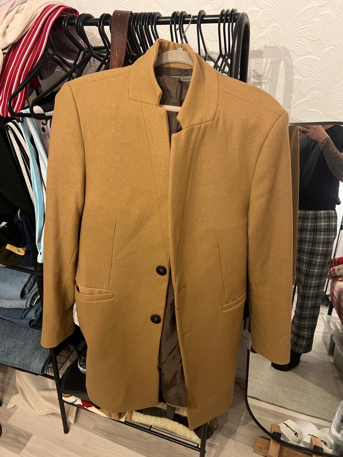 Camel coat hanging on a clothes rail with other garments. Reflection of a person in the mirror in the background. Shop environment.