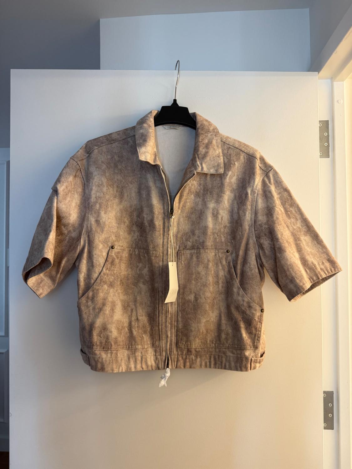 Brown-toned, short-sleeved jacket hanging on a black hanger over a white door. It has a zip and front pockets.