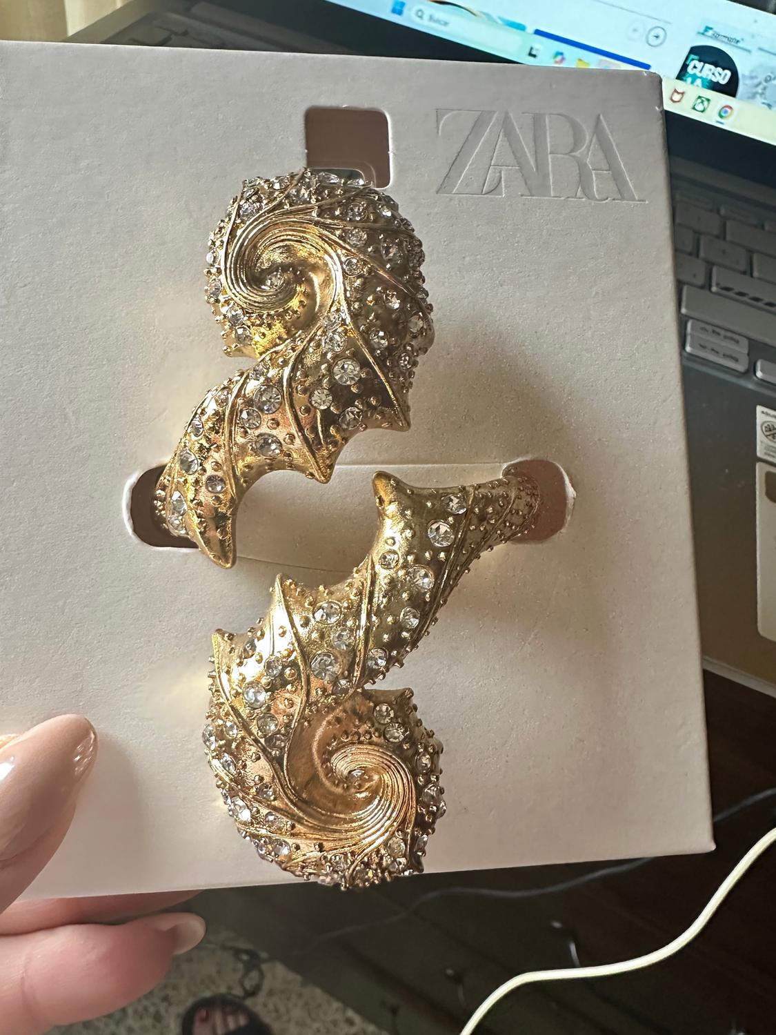 Gold brooch with brilliant spiral-shaped inlays on white cardboard with Zara logo, laptop background.