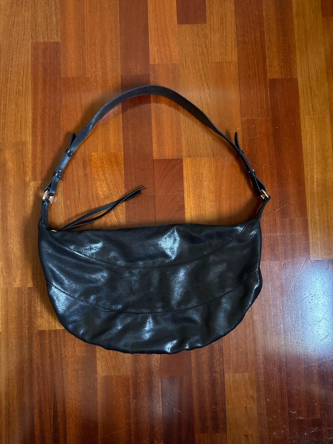 Black crescent-shaped shoulder bag with a gloss finish and an adjustable strap, resting on a wooden floor.